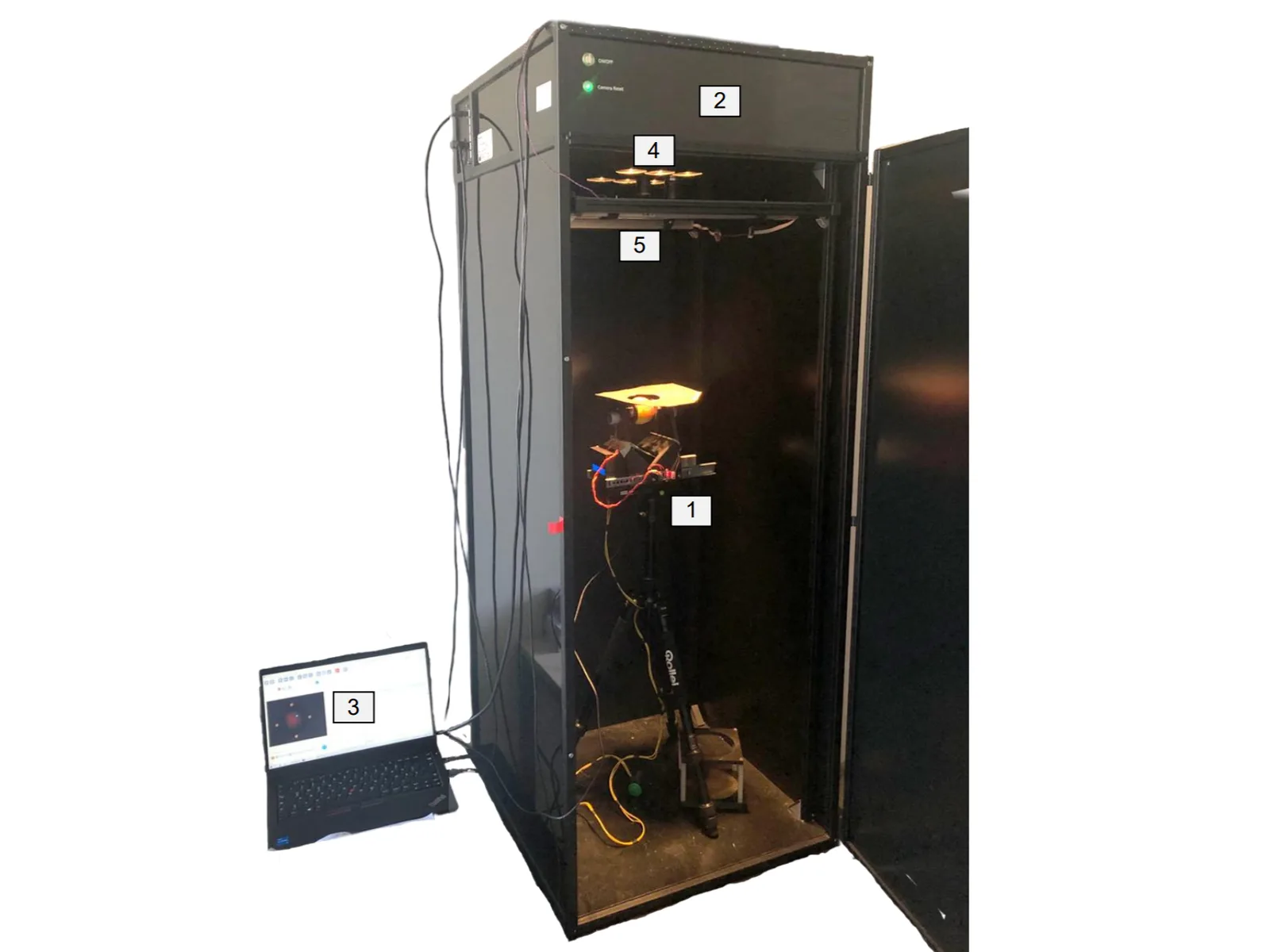 A black hyperspectral imaging cabinet with its door open: lamps at the top light the rotator standing on a tripod inside, and a laptop beside the cabinet shows the camera view of the apple. Numbered labels mark the parts.