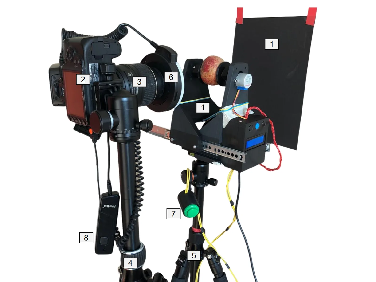 The rotator on a tripod, holding an apple between two black 3D-printed arms with a small stepper motor on one arm and a control box with display and buttons below; a DSLR with a ring light on a second tripod faces the apple against a black backdrop. Numbered labels mark the parts.